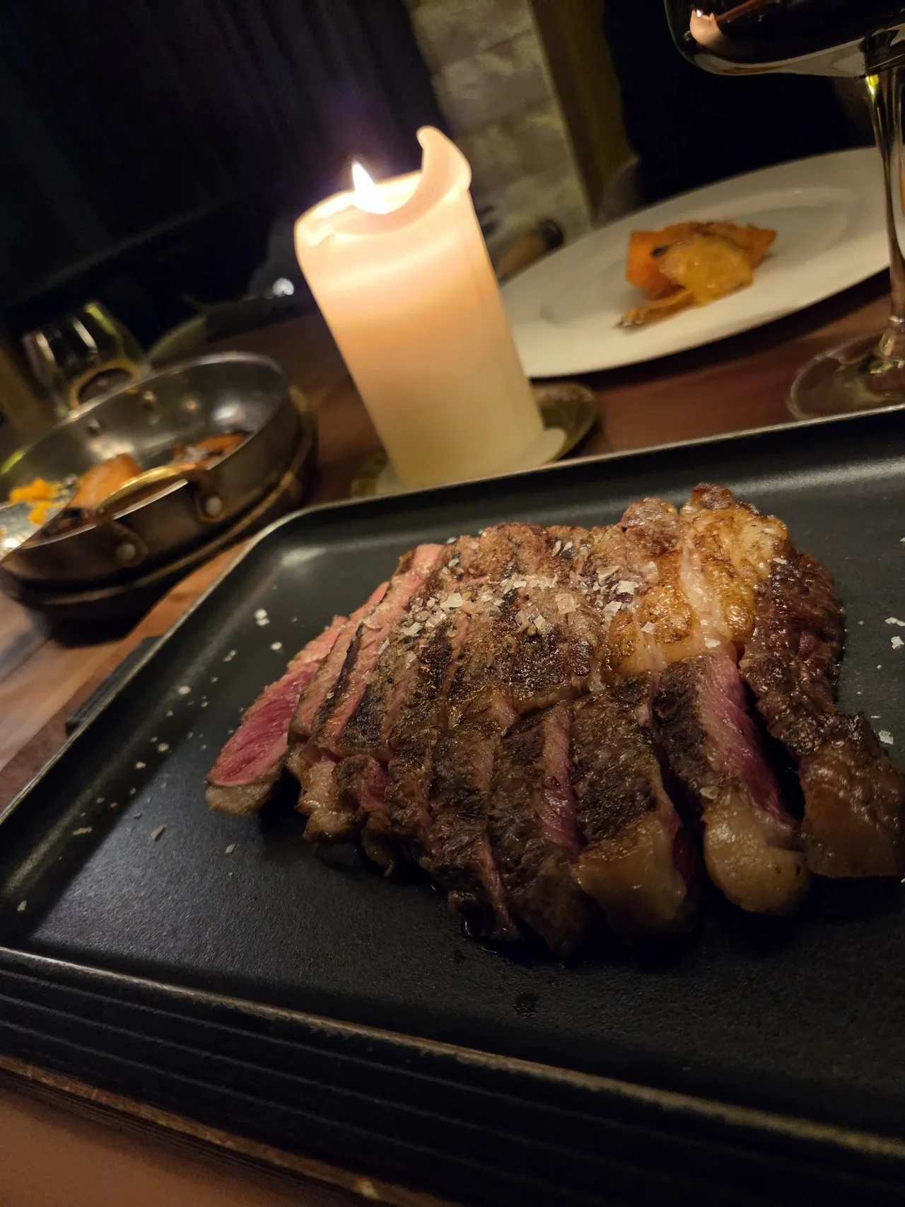 A sliced grilled steak on a dark slate plate beside a candle
