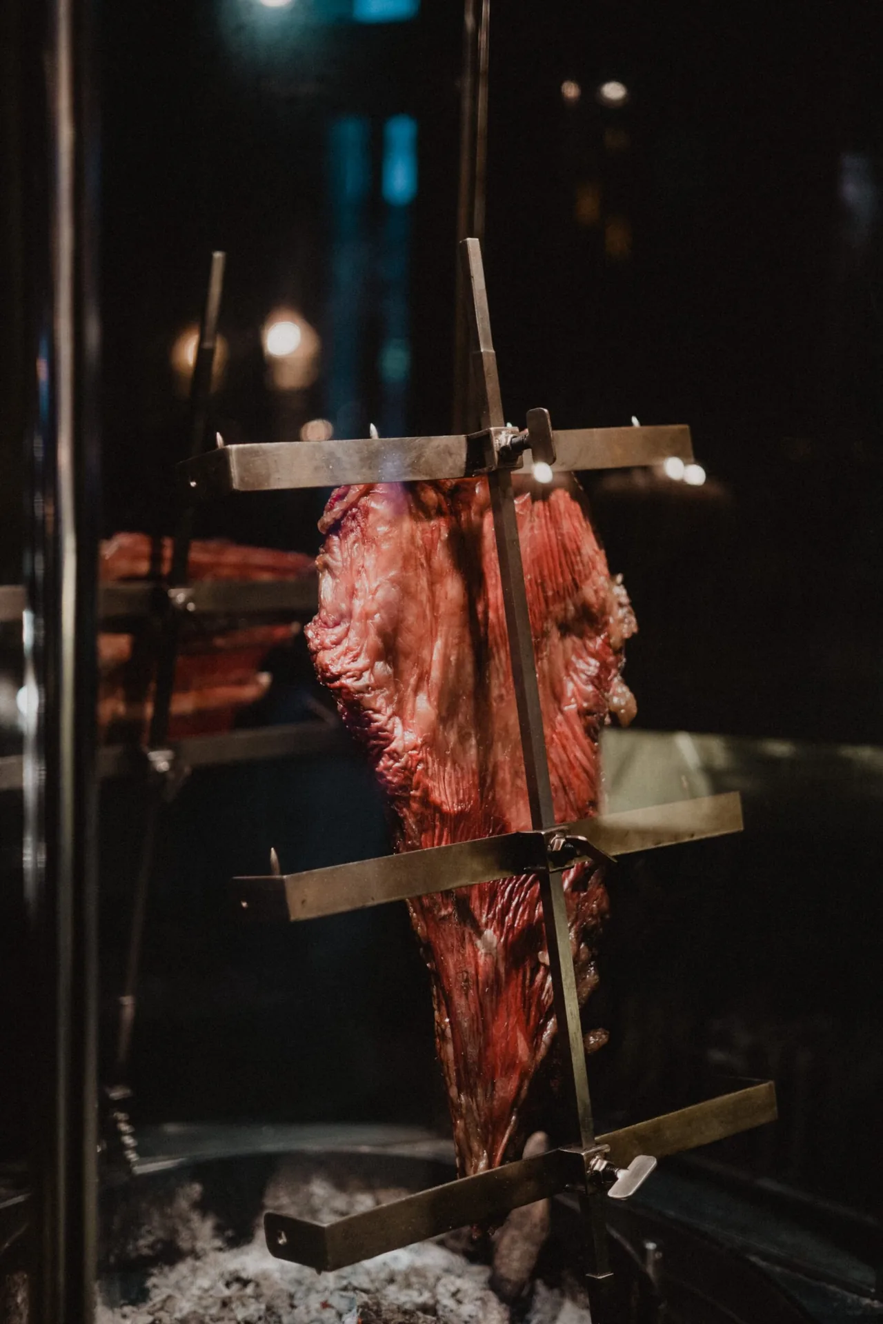 A large cut of beef roasting on an upright metal cross over embers