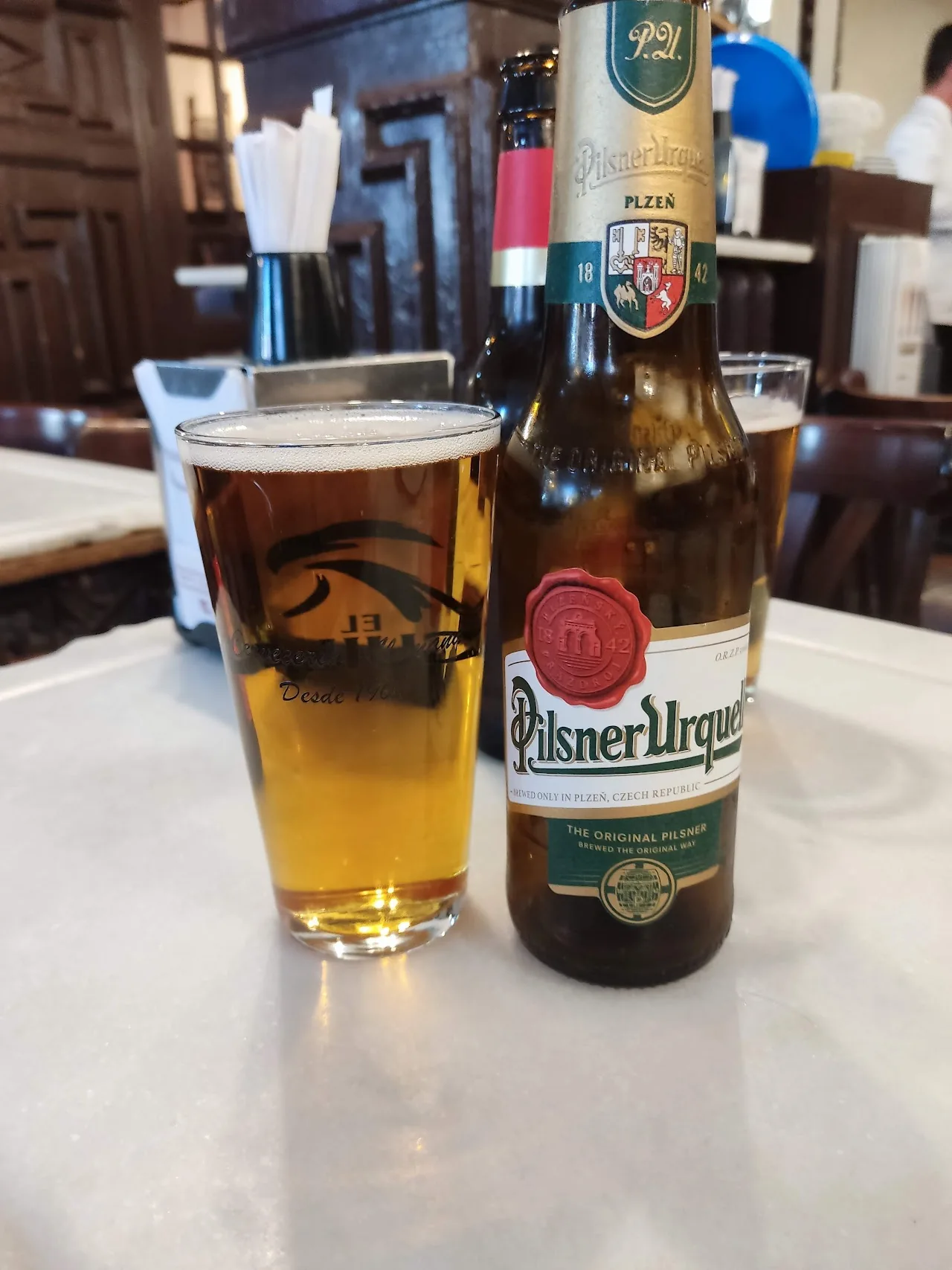 A glass of draft beer next to a bottle of Pilsner Urquell on a table