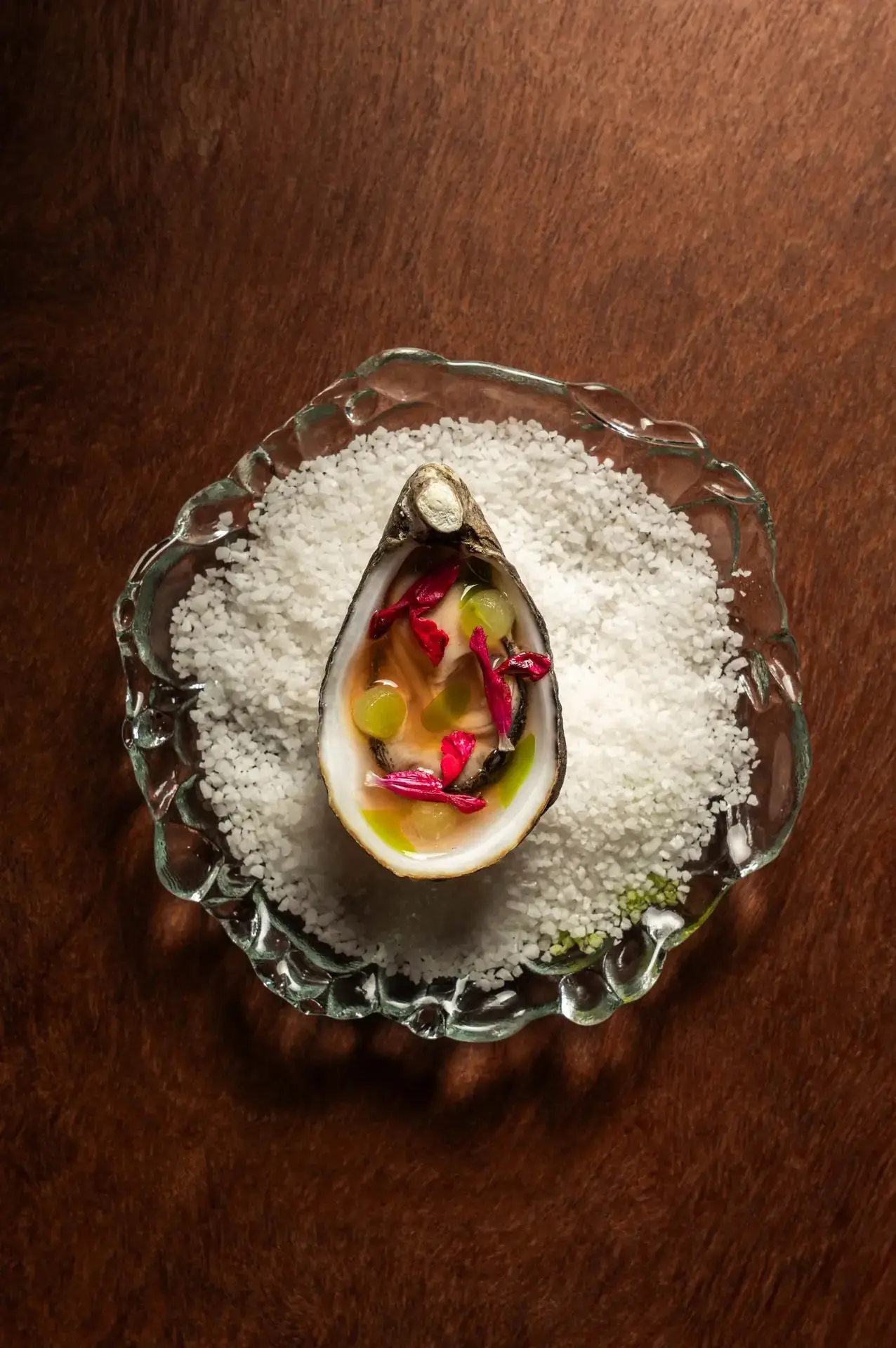 Half oyster on the half shell topped with grapes, a savory glaze, and red flower petals, served over rock salt on a scalloped glass dish, at Chambacú