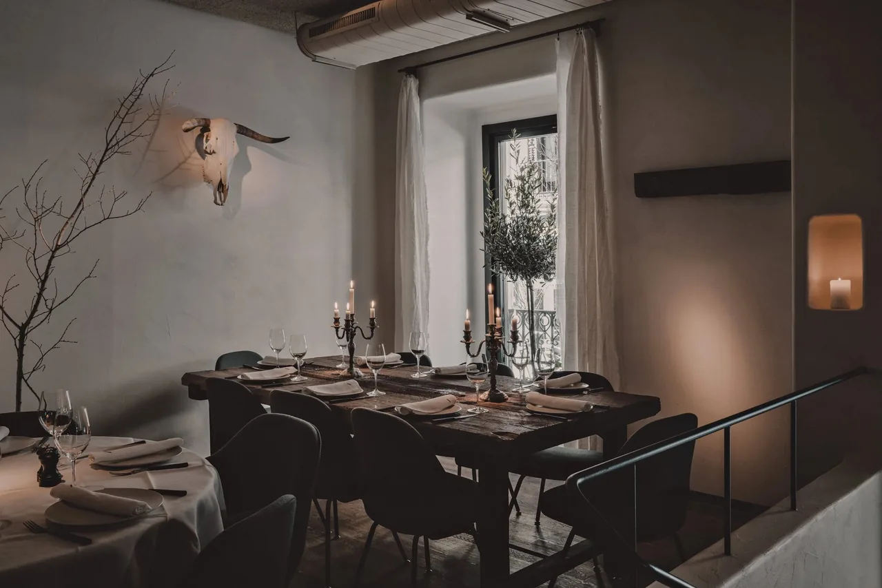 A dim dining room with set tables, candles, bare branches and a large window