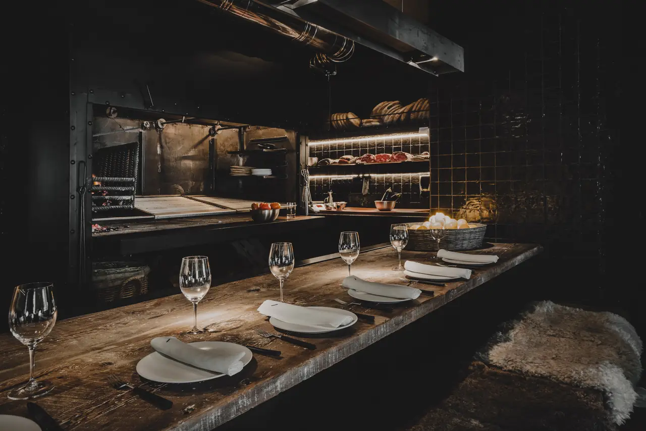 Set dining table with wine glasses and folded napkins beside the open charcoal grill and a meat display shelf at Charrúa
