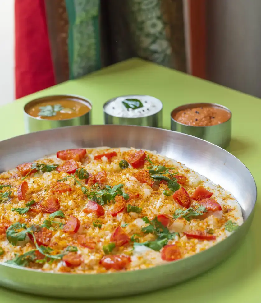 A masala uttapam topped with diced tomatoes, green chilies, and chopped cilantro, served on a round steel plate alongside three metal cups of sambar, coconut chutney, and tomato chutney, Chennai Masala Dosa
