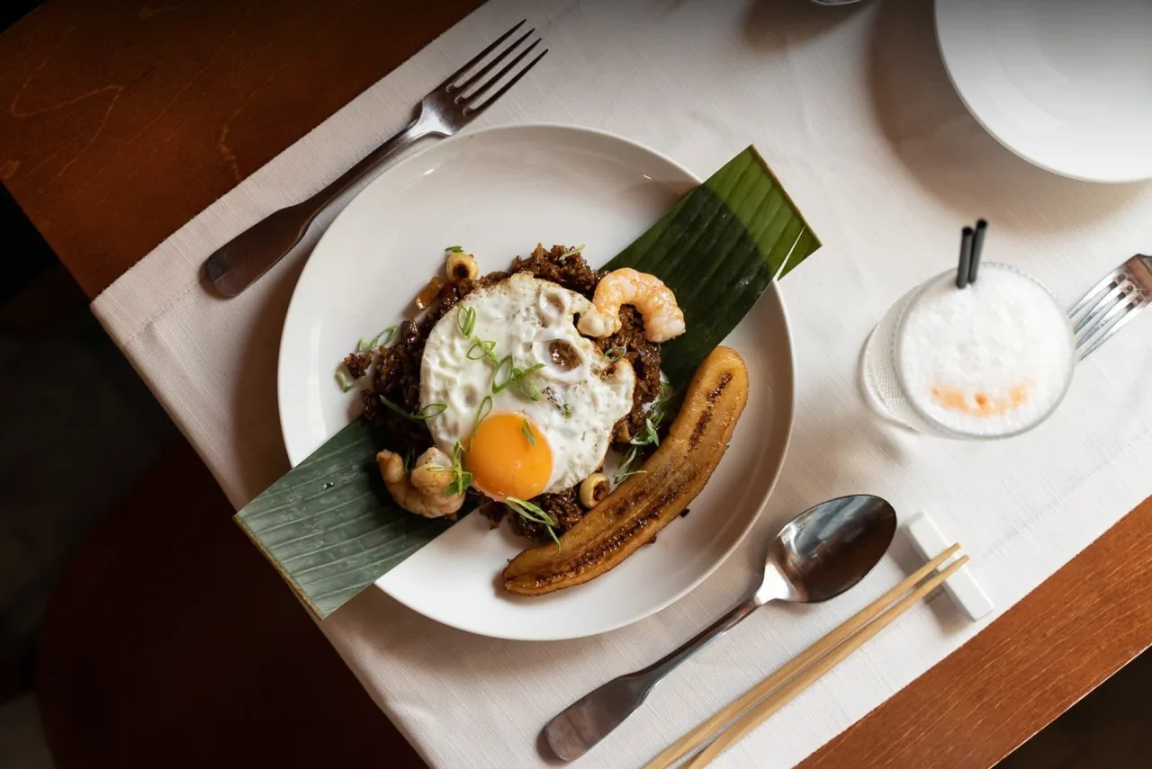 A plate of dark fried rice topped with a fried egg and a fried banana slice on a banana leaf