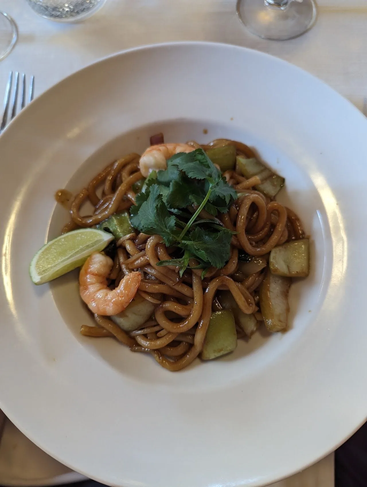 A plate of stir-fried noodles with prawns, vegetables and herbs and a lime wedge