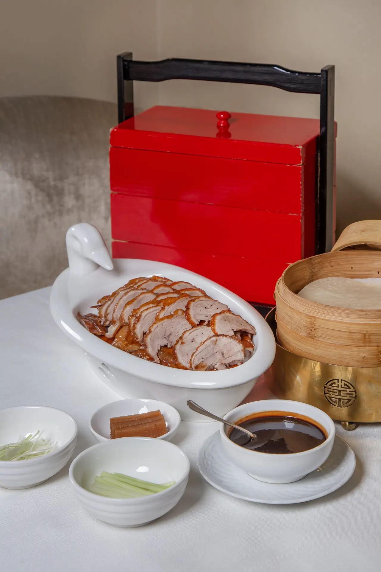 Sliced Peking duck arranged in a white duck-shaped dish, served with a stacked bamboo steamer of pancakes, bowls of shredded scallion and cucumber, and a bowl of dark hoisin sauce, a red lacquered box behind