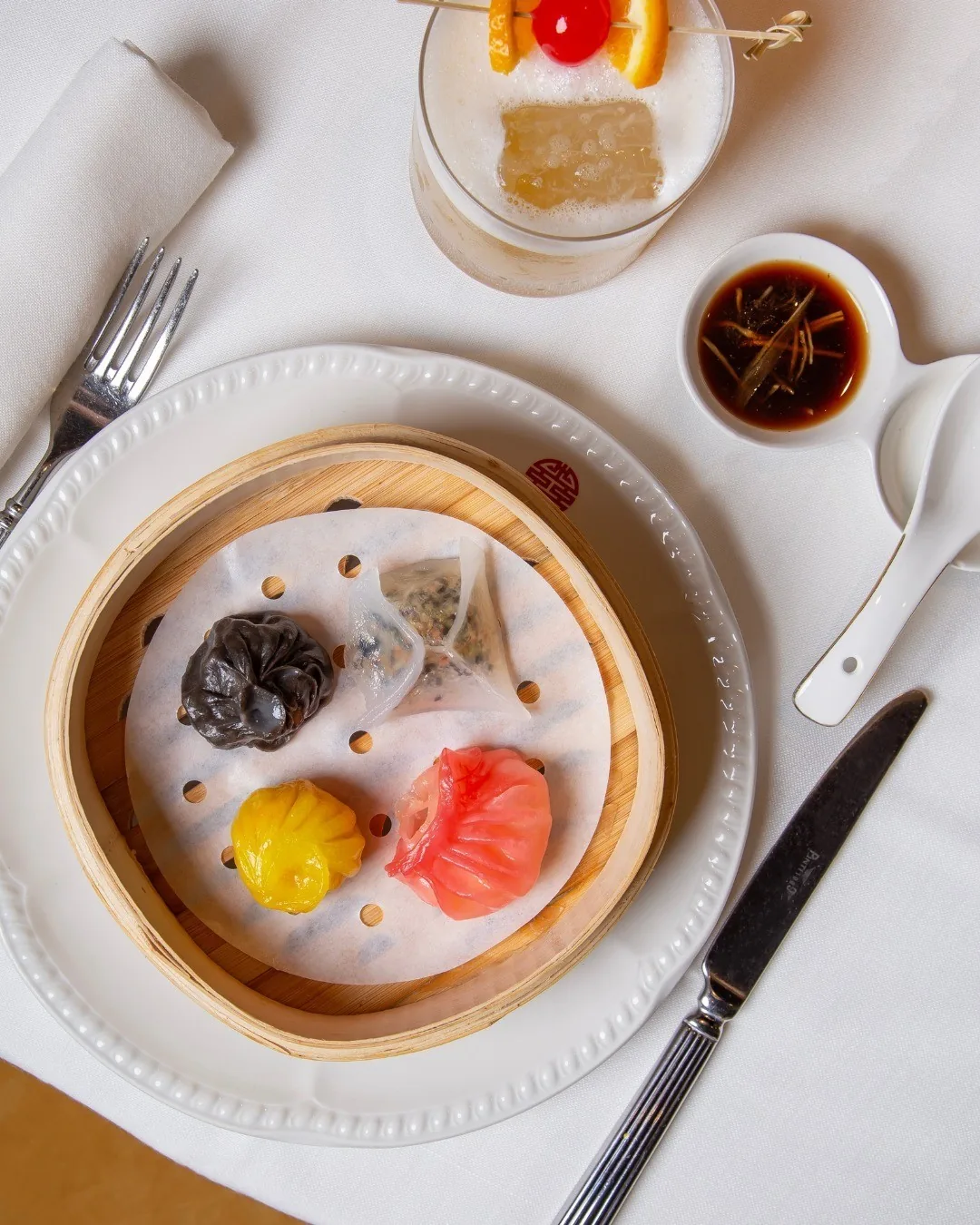 A bamboo steamer of assorted dumplings on a white plate with a dipping sauce beside it