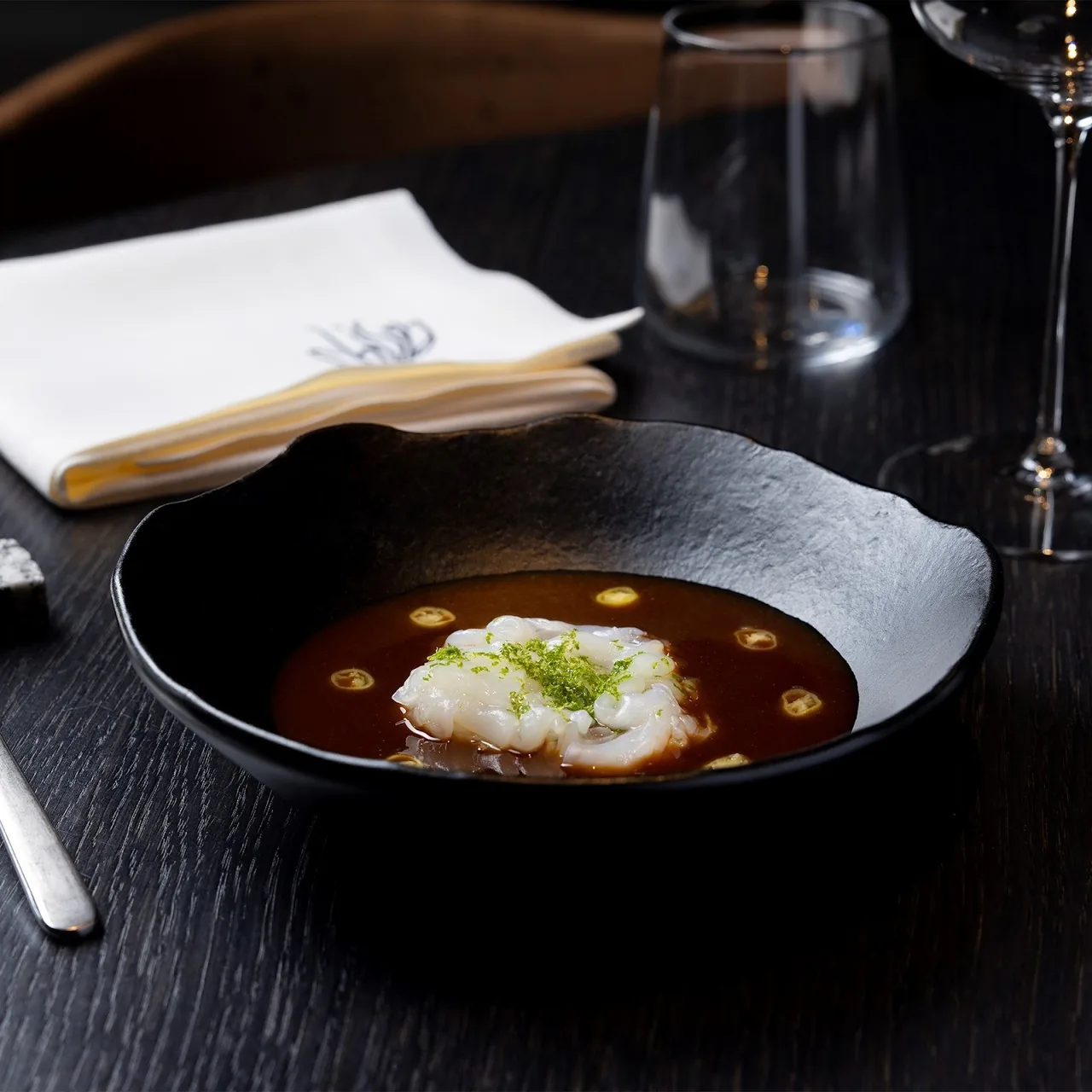 A black bowl of dark broth with a white fish fillet and green dots