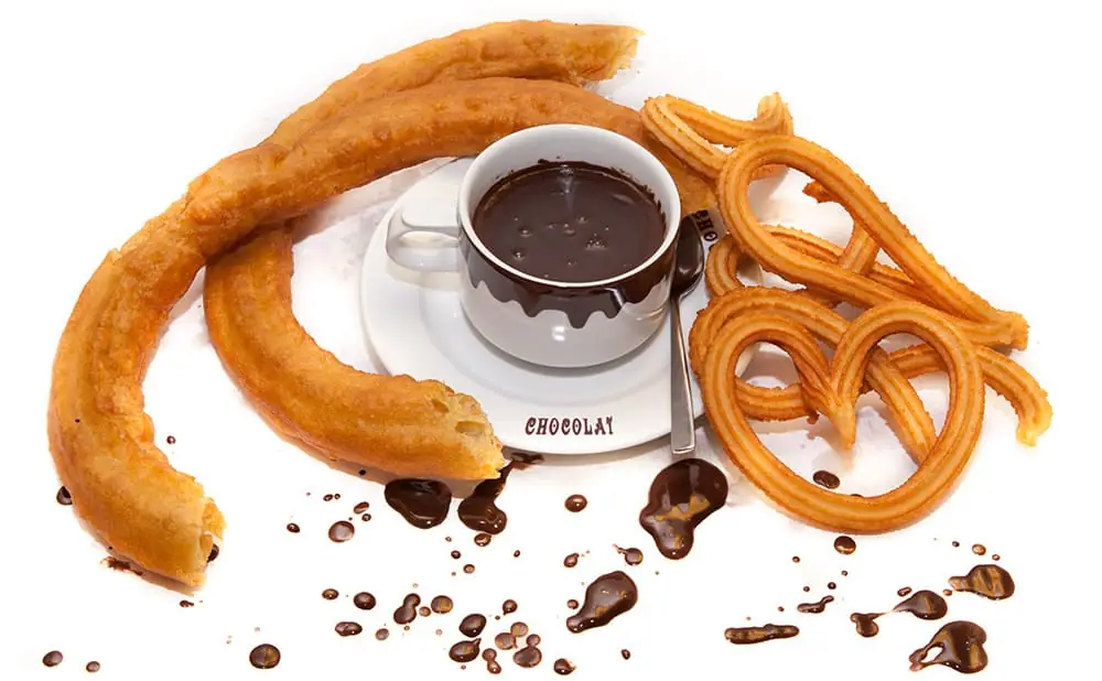 Churros and porras arranged around a cup of thick hot chocolate with chocolate splatters on a white background
