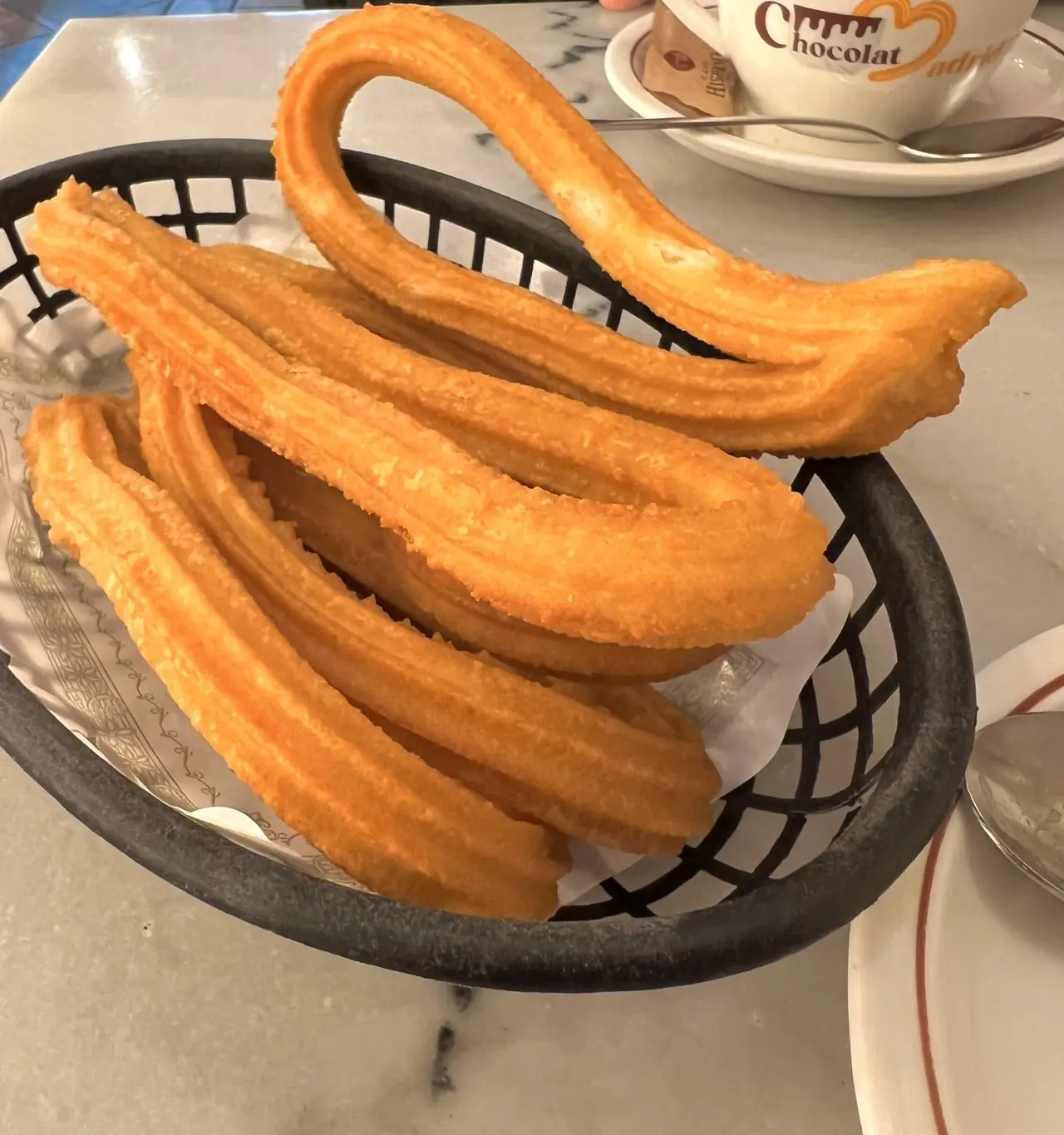 Long looped churros in a black basket, with a branded cup of hot chocolate behind