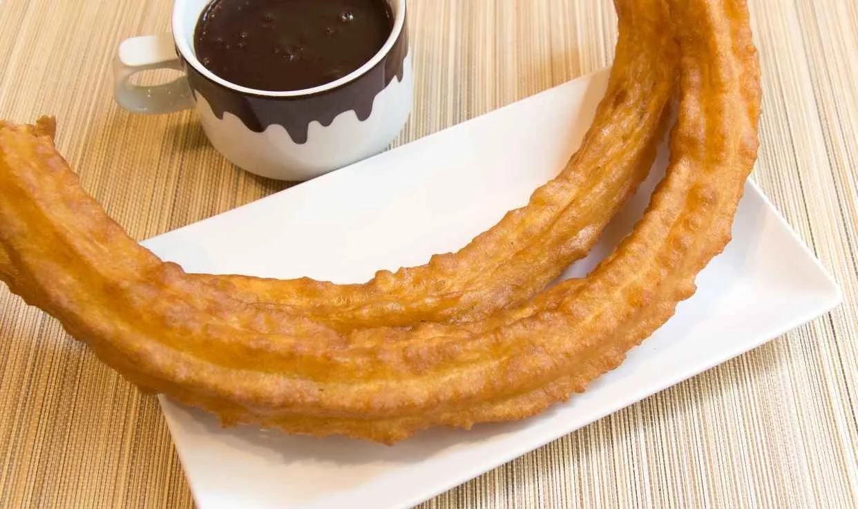 A large coiled churro on a white plate with a cup of chocolate behind it