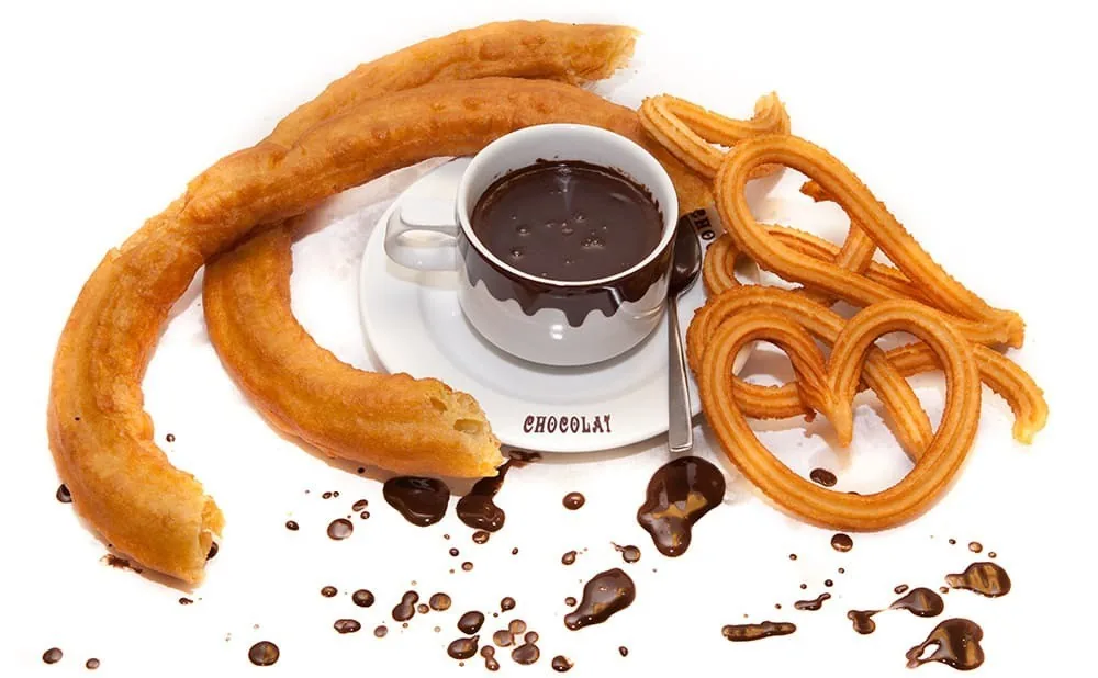 A pile of churros and a cup of hot chocolate with chocolate crumbs on a white background