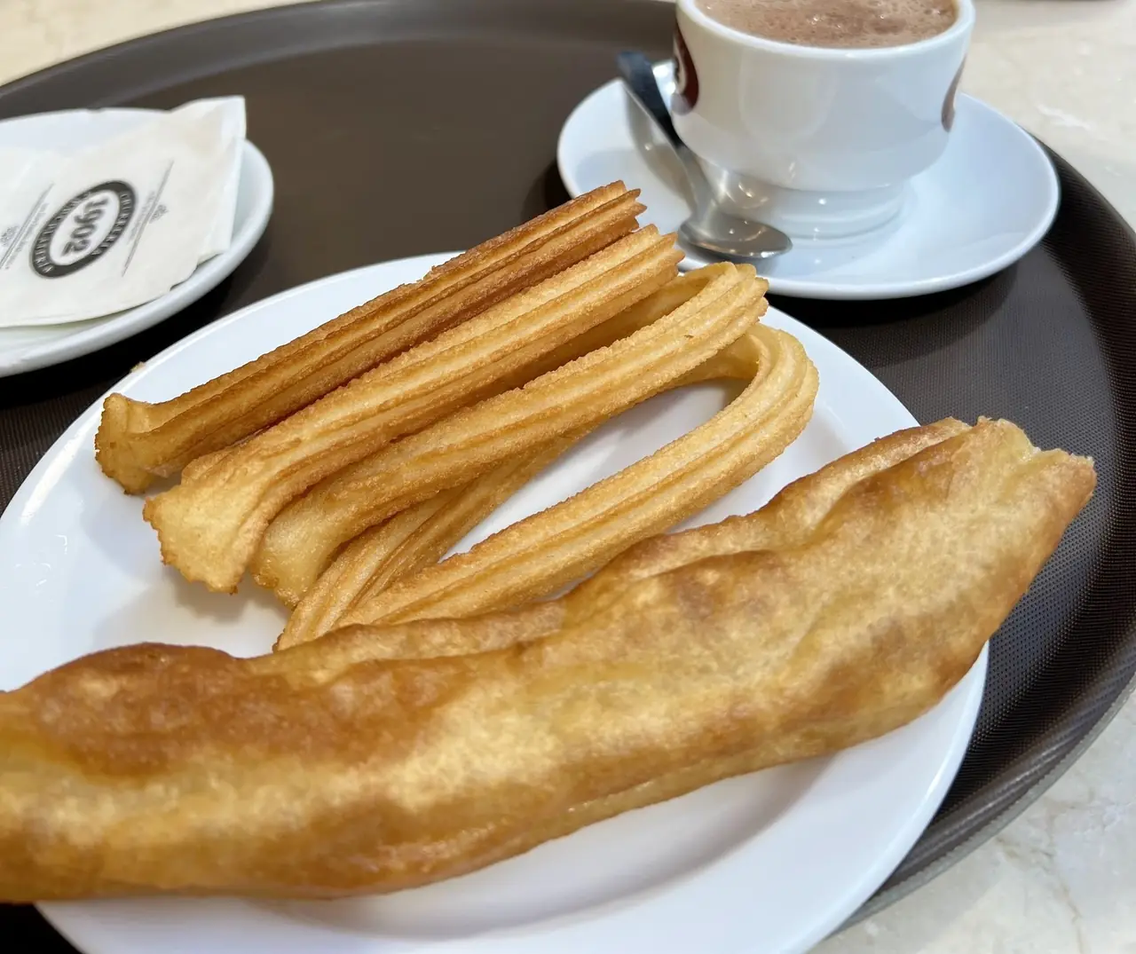 Thin churros and a thick porra on a white plate, with a cup of hot chocolate behind
