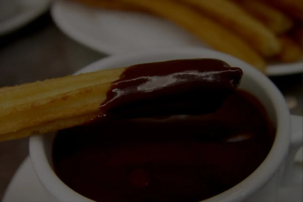 Golden churro dipped into a white cup of thick dark hot chocolate, with more churros on a plate behind