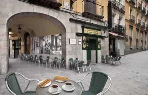 Outdoor terrace under a stone arcade with a plate of churros and two cups of chocolate on a foreground table, Chocolateria San Gines signage visible across the plaza