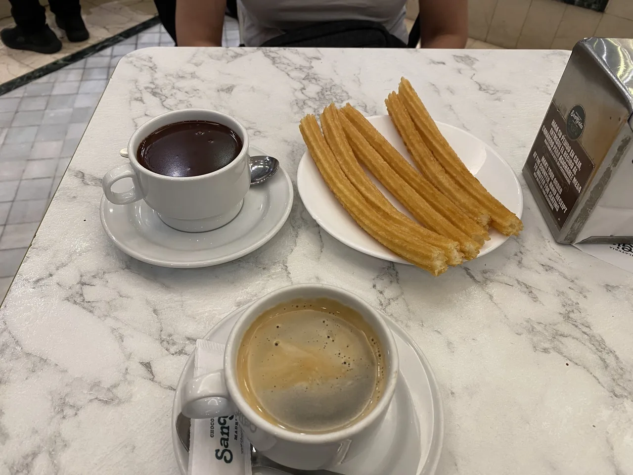 A cup of hot chocolate and a cup of coffee with a plate of churros on a marble table