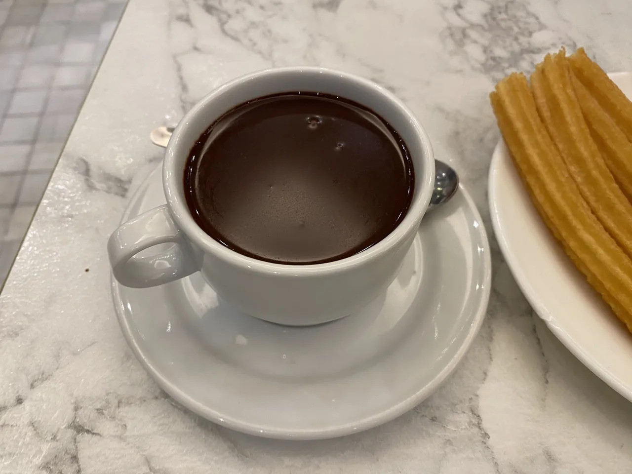 A white cup of thick hot chocolate beside a plate of churros on a marble table