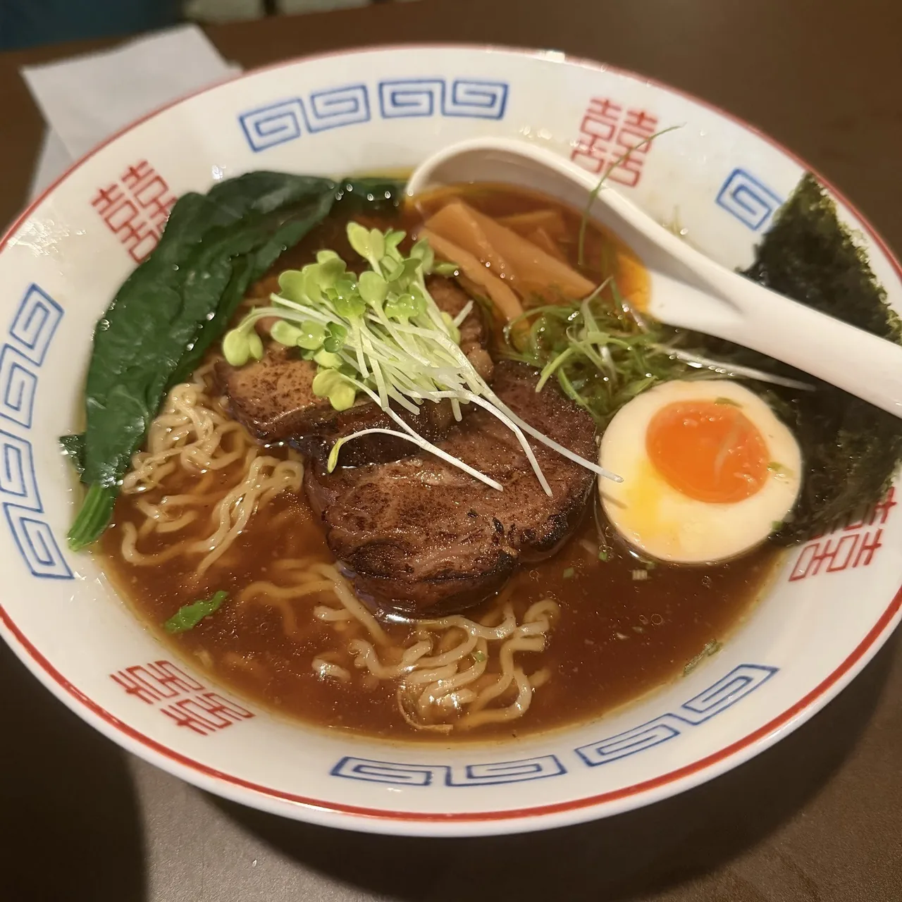 A bowl of ramen in dark broth with slices of pork, a halved egg, seaweed and a white spoon, seen from above