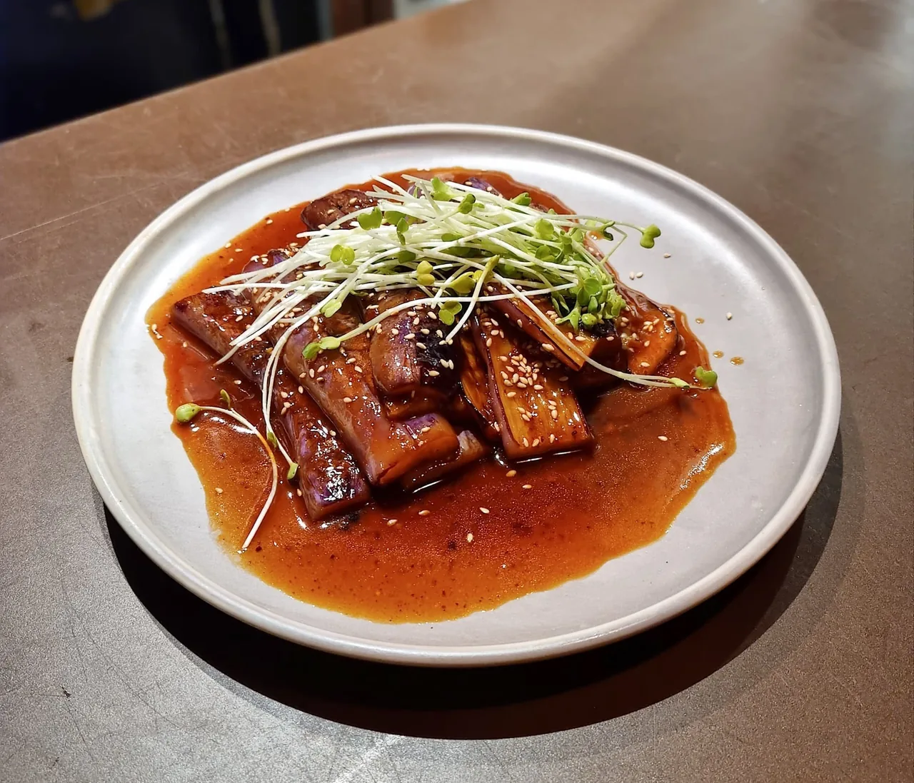 Glazed dark pieces of fish topped with green shoots in red-brown sauce on a round grey plate