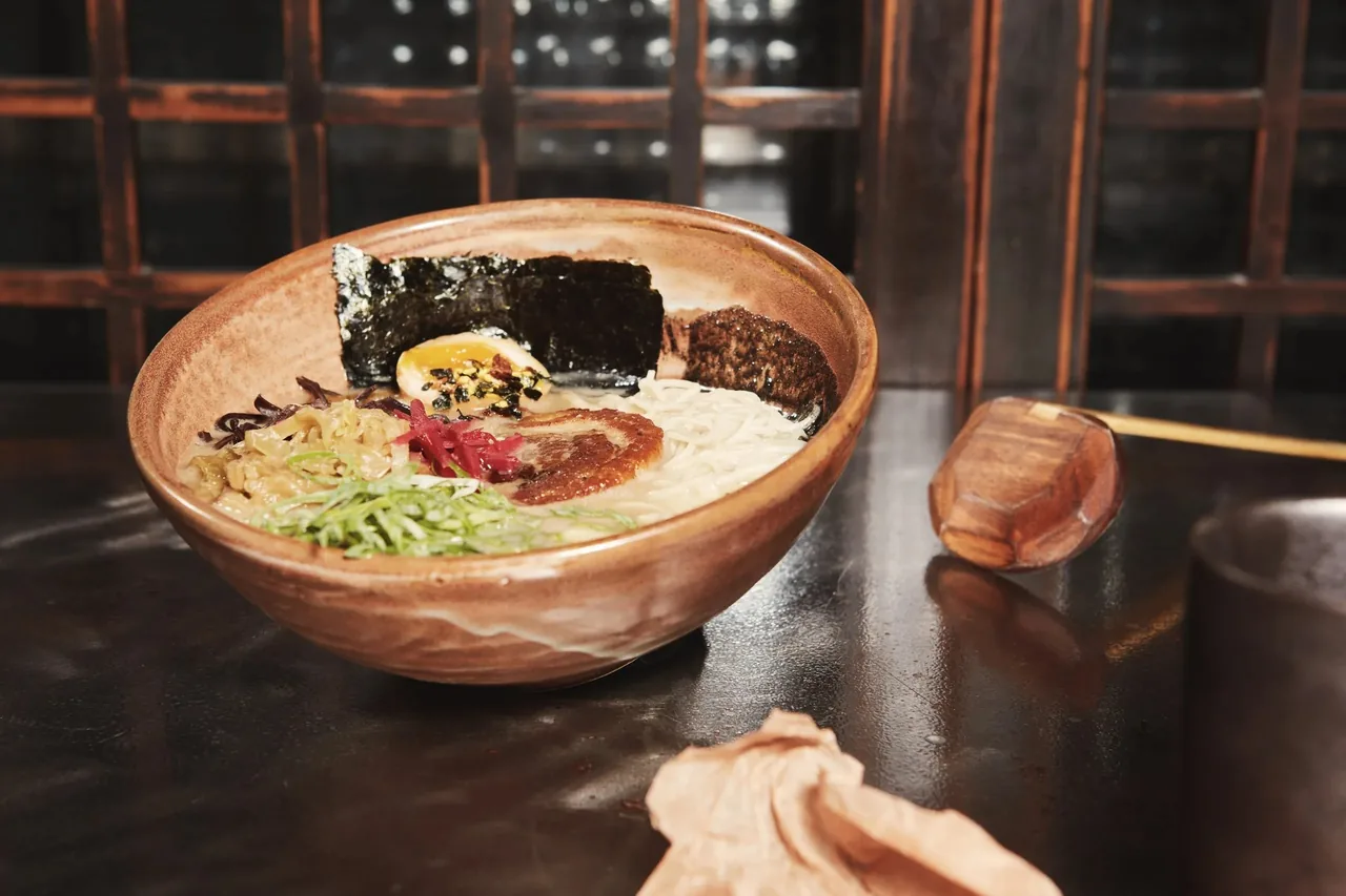 A ramen bowl with noodles, seaweed, a halved egg and spring onion in a terracotta-coloured bowl on a dark table