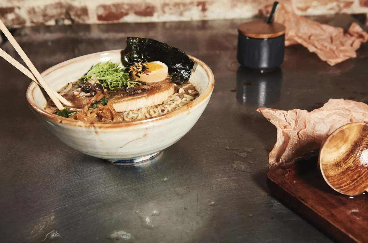 A bowl of ramen with a slice of pork, a halved egg, seaweed and mushrooms with chopsticks resting in the bowl