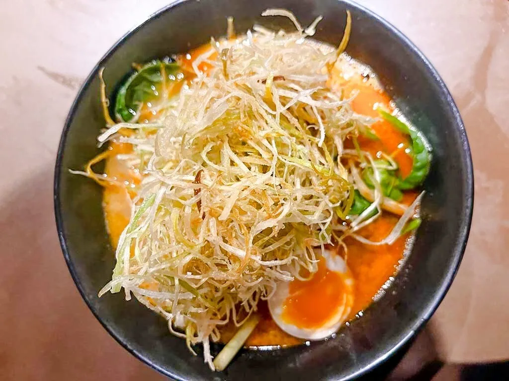 A dark bowl of orange broth topped with a pile of fried shredded leek and a halved egg