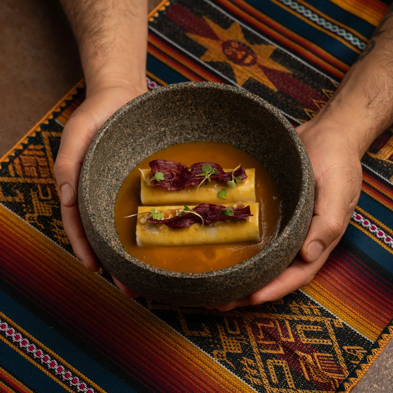Two hands cradling a rough stone bowl holding two pale cannelloni in a russet broth, topped with dark red root-vegetable crisps and micro leaves, set on a striped Andean textile