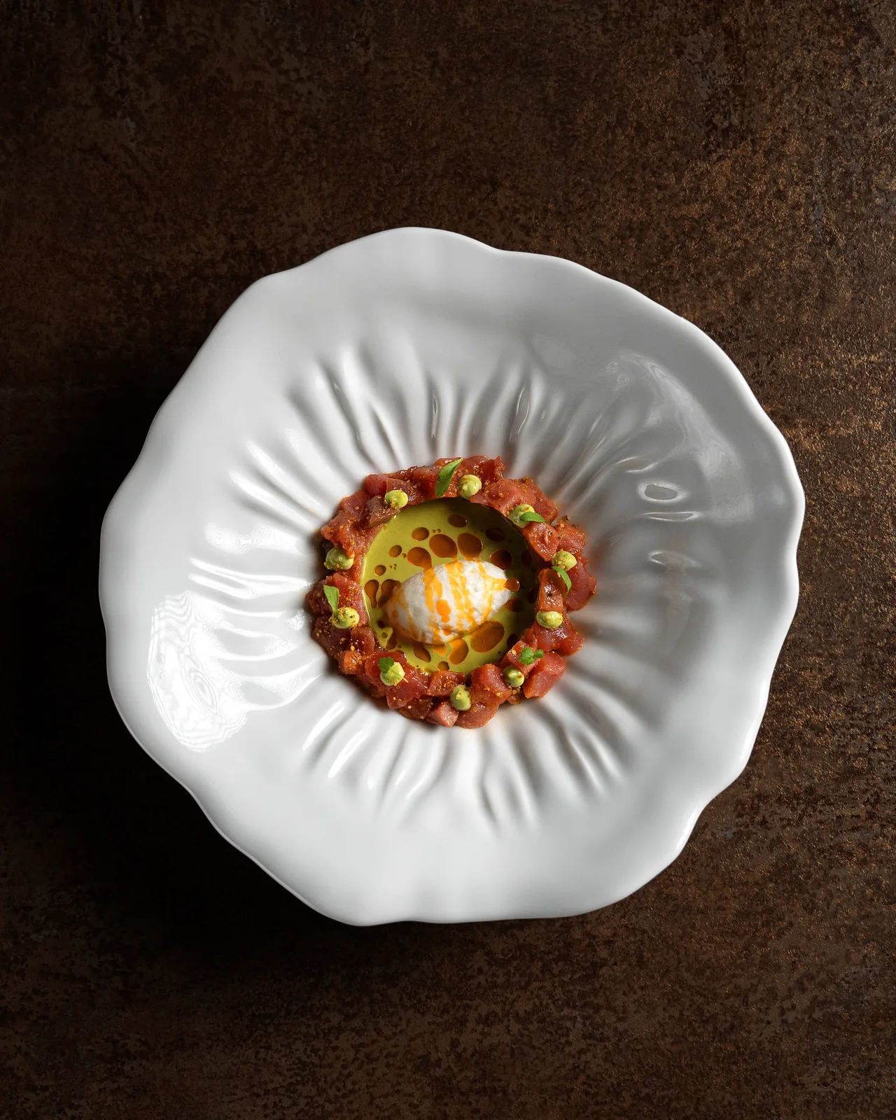 A ring of diced raw red fish studded with green dots and micro leaves, encircling a pool of bright green sauce with a white quenelle drizzled orange, on a sculpted white plate against a dark rust background