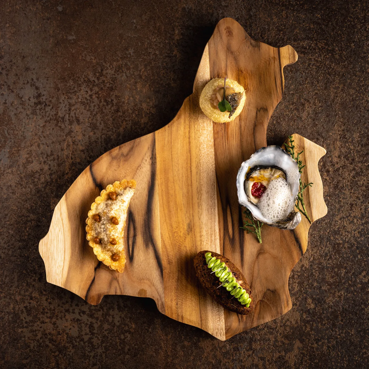 A shaped wooden board carrying four canapes: a crimped fried empanada dusted with cheese, a crisp potato nest, an oyster on the half shell under a light foam with a sprig of greenery, and a dark bread base piped with green sauce
