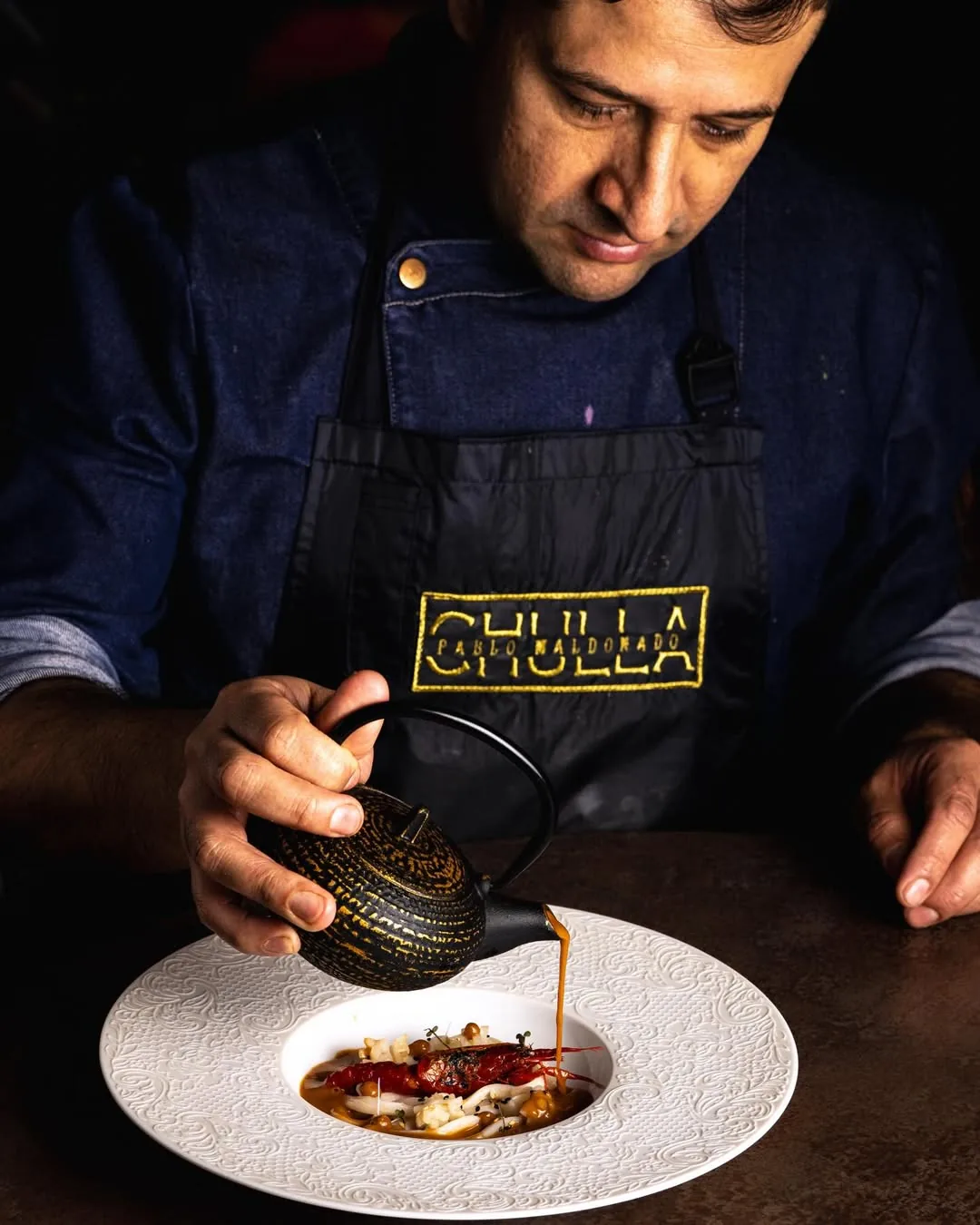 A chef in a CHULLA-branded apron pouring a dark broth from a cast-iron teapot onto a plate of charred red peppers, shellfish and diced garnish on an embossed white plate