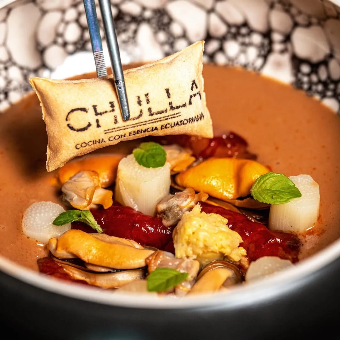A deep bowl of shellfish in a pale russet broth with clams, scallop-white discs, charred red pepper and mint leaves, with a thin wafer stamped CHULLA - COCINA CON ESENCIA ECUATORIANA held above it by tweezers