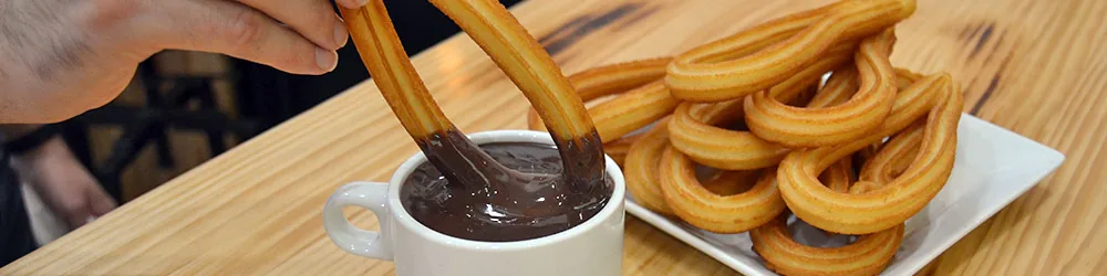 A hand dipping two long churros into a cup of thick dark drinking chocolate, with a white plate of looped churros alongside on a pale wooden table