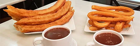 Two white plates on a pale counter, one of thick ridged porras and one of looped churros, each with a white cup of thick dark chocolate in front
