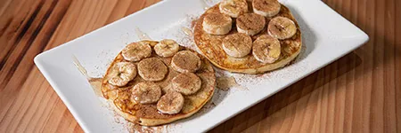 Two round pancakes on a white rectangular plate, each fanned with banana slices and dusted with cinnamon, on a wooden table