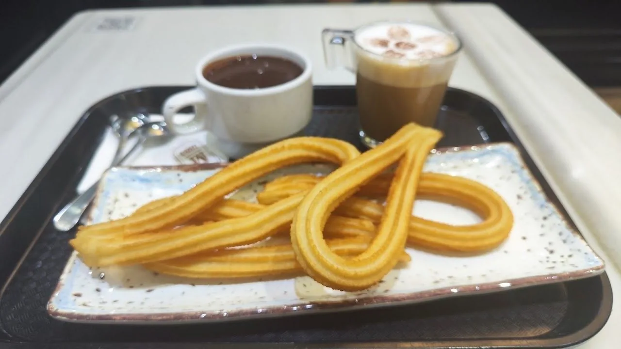 Looped churros on a blue and white plate with a cup of chocolate and a coffee on a tray