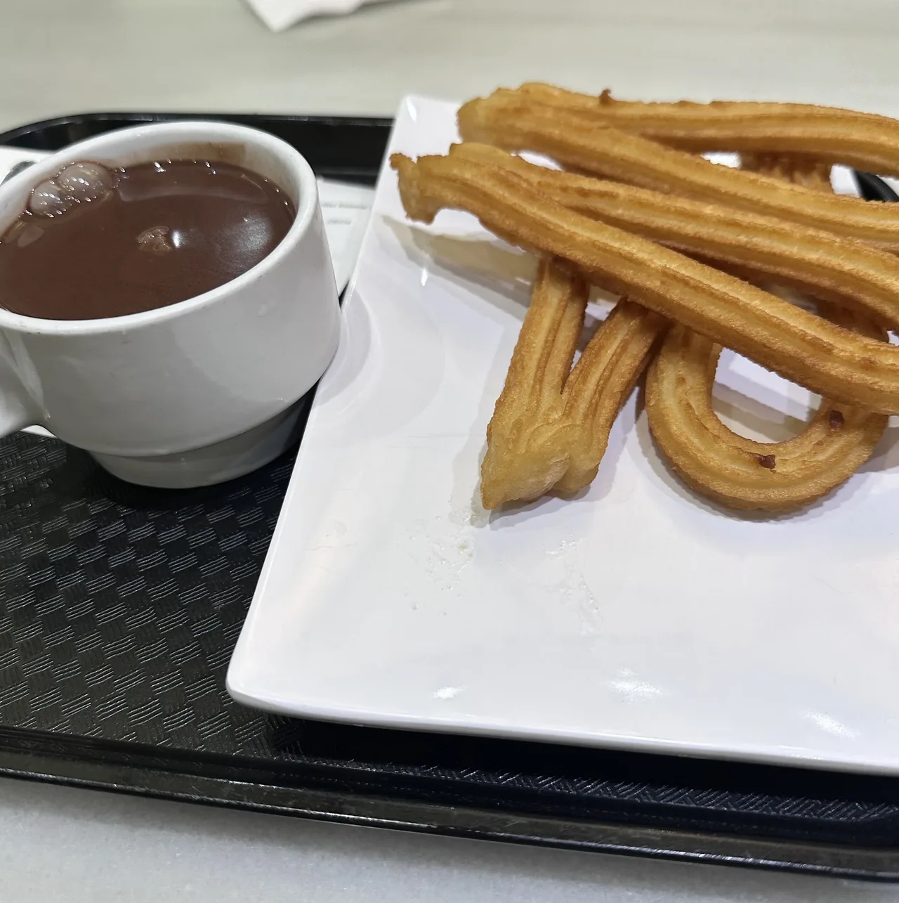A cup of hot chocolate and a plate of golden churros on a black tray