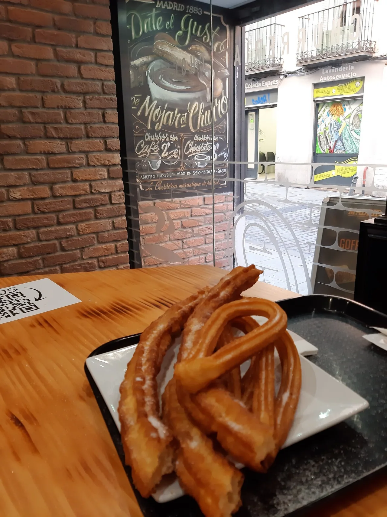 A plate of sugared churros on a wooden table beside a brick wall