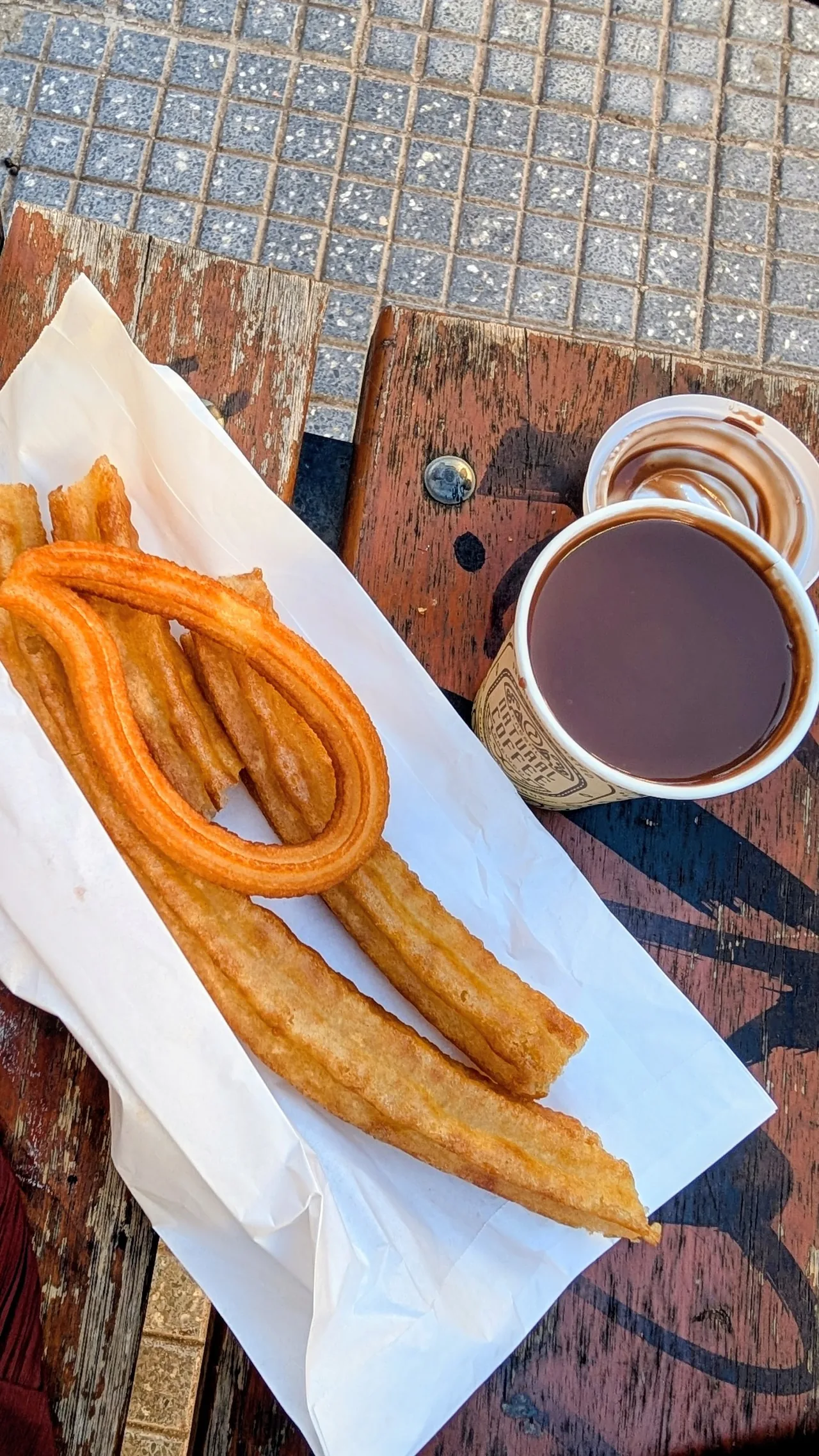 Churros on white paper beside a cup of hot chocolate on a wooden bench