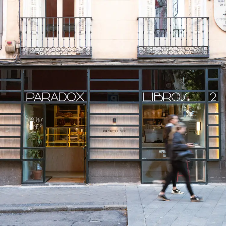 Street corner exterior beneath 'PARADOX' and 'LIBROS' signs with the 'CIENTOTREINTA' bakery window and its bread display, passersby blurred in front