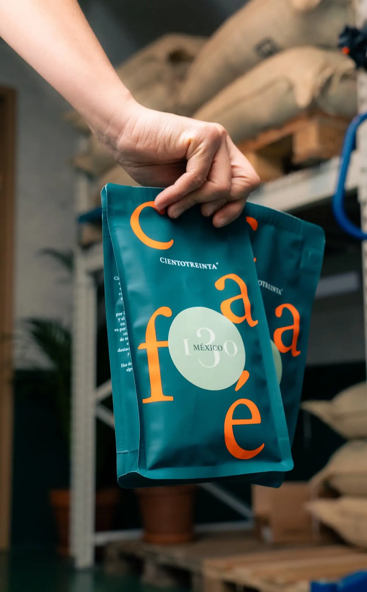 Hand holding up two teal Cientotreinta 'Mexico' coffee bags in the roastery, burlap sacks of green coffee stacked behind