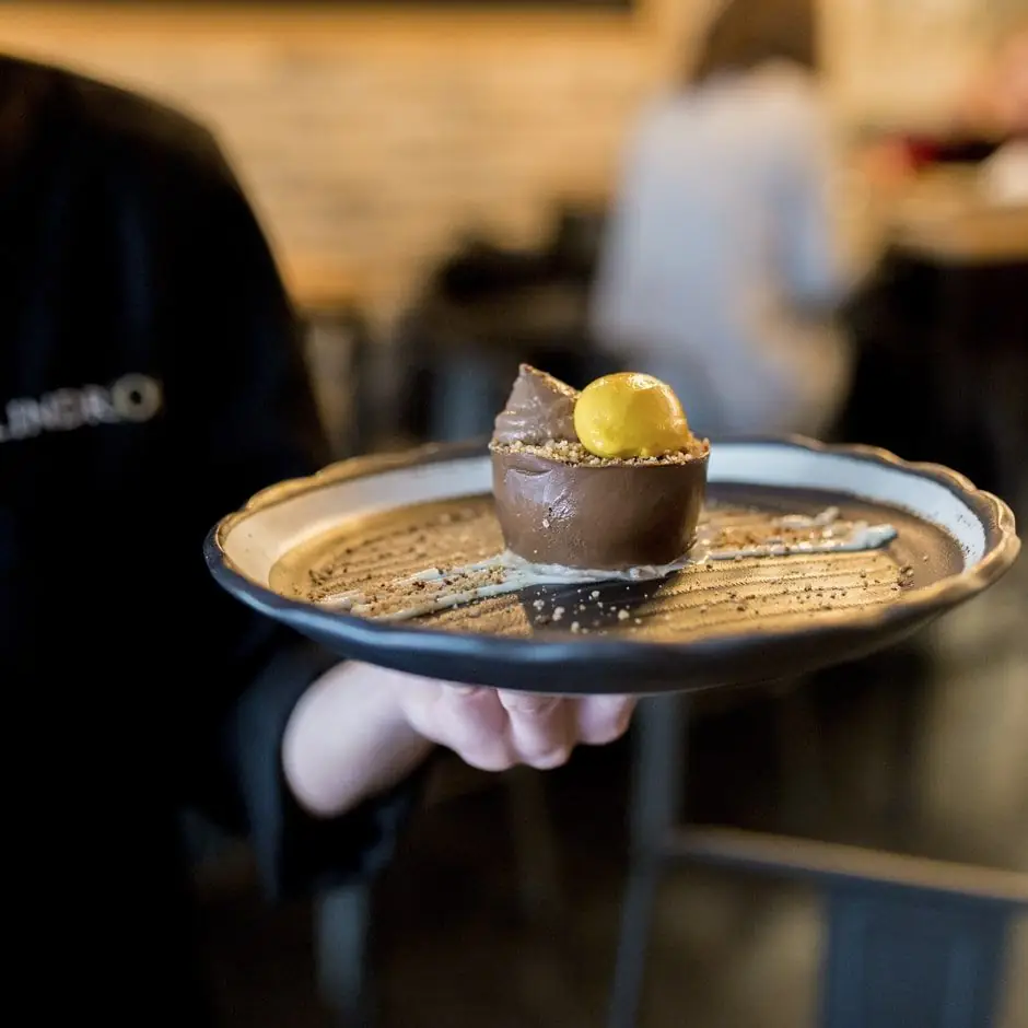 Chocolate mousse dome topped with a yellow sorbet quenelle on a black scalloped plate, held by staff wearing a Cilindro-branded apron
