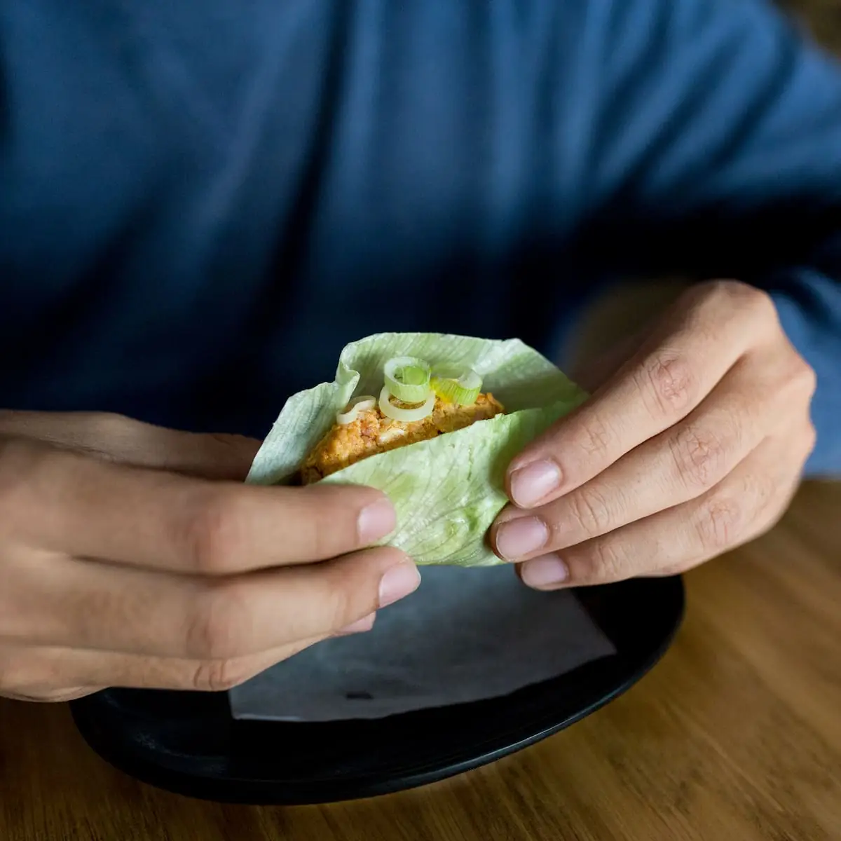 Hands holding a lettuce wrap filled with an orange fritter and topped with sliced scallion, over a black plate