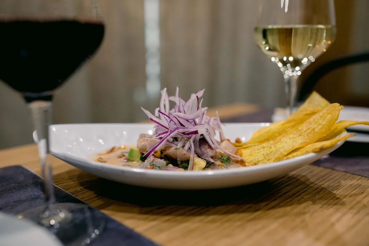 A white oval bowl of sliced fish in a creamy sauce with red onion and fried plantain chips beside a glass of wine