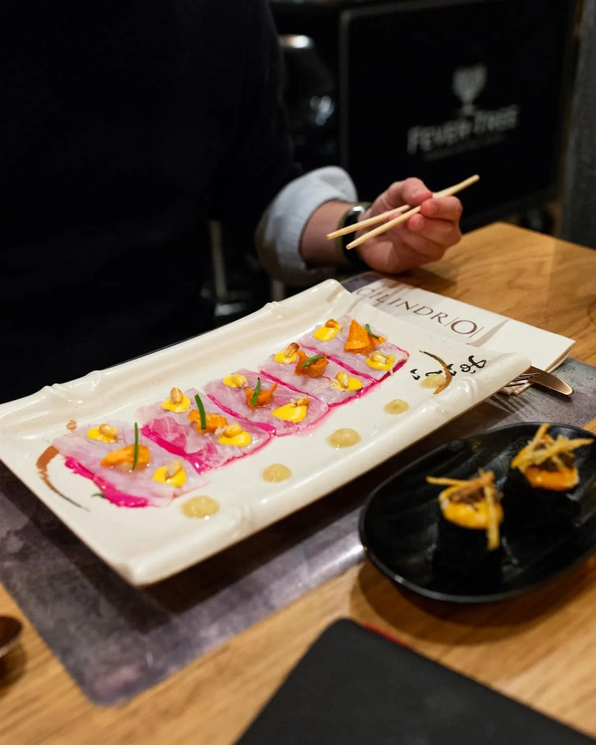 A rectangular plate of sliced raw fish with pink and yellow garnishes and a hand holding chopsticks