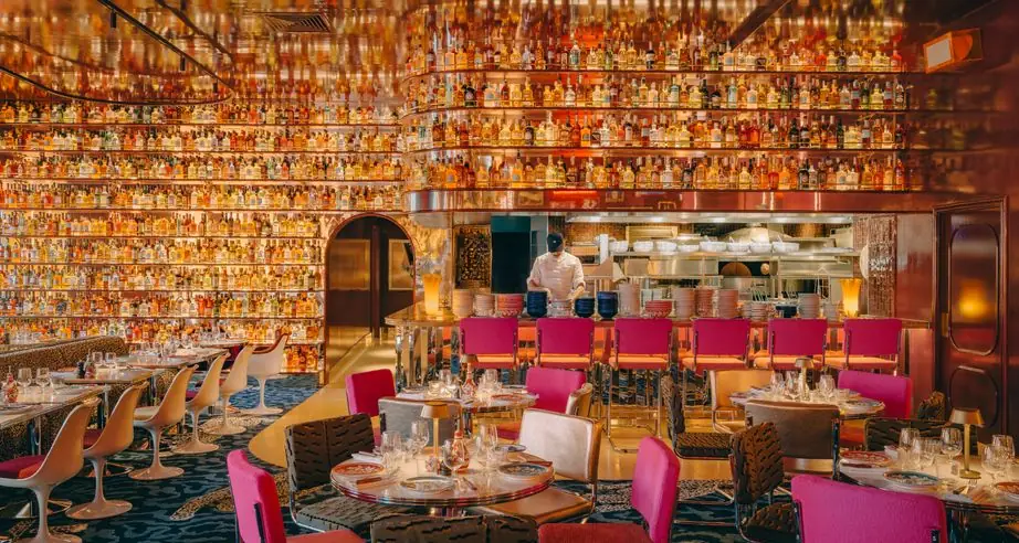 Dramatic dining room with curved walls lined floor-to-ceiling with backlit liquor bottles, hot-pink and animal-print chairs, round tables and an open kitchen pass