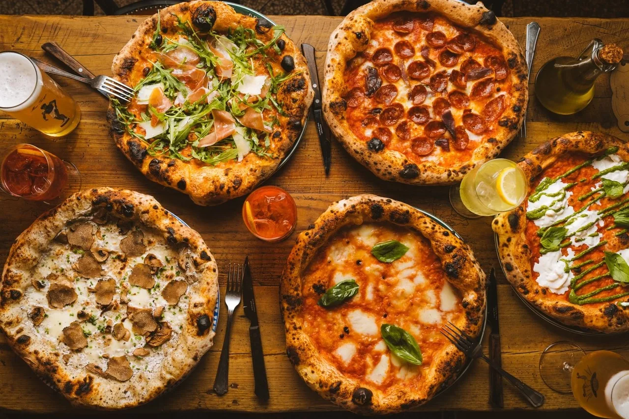 Four pizzas with different toppings on a wooden table with drinks and cutlery