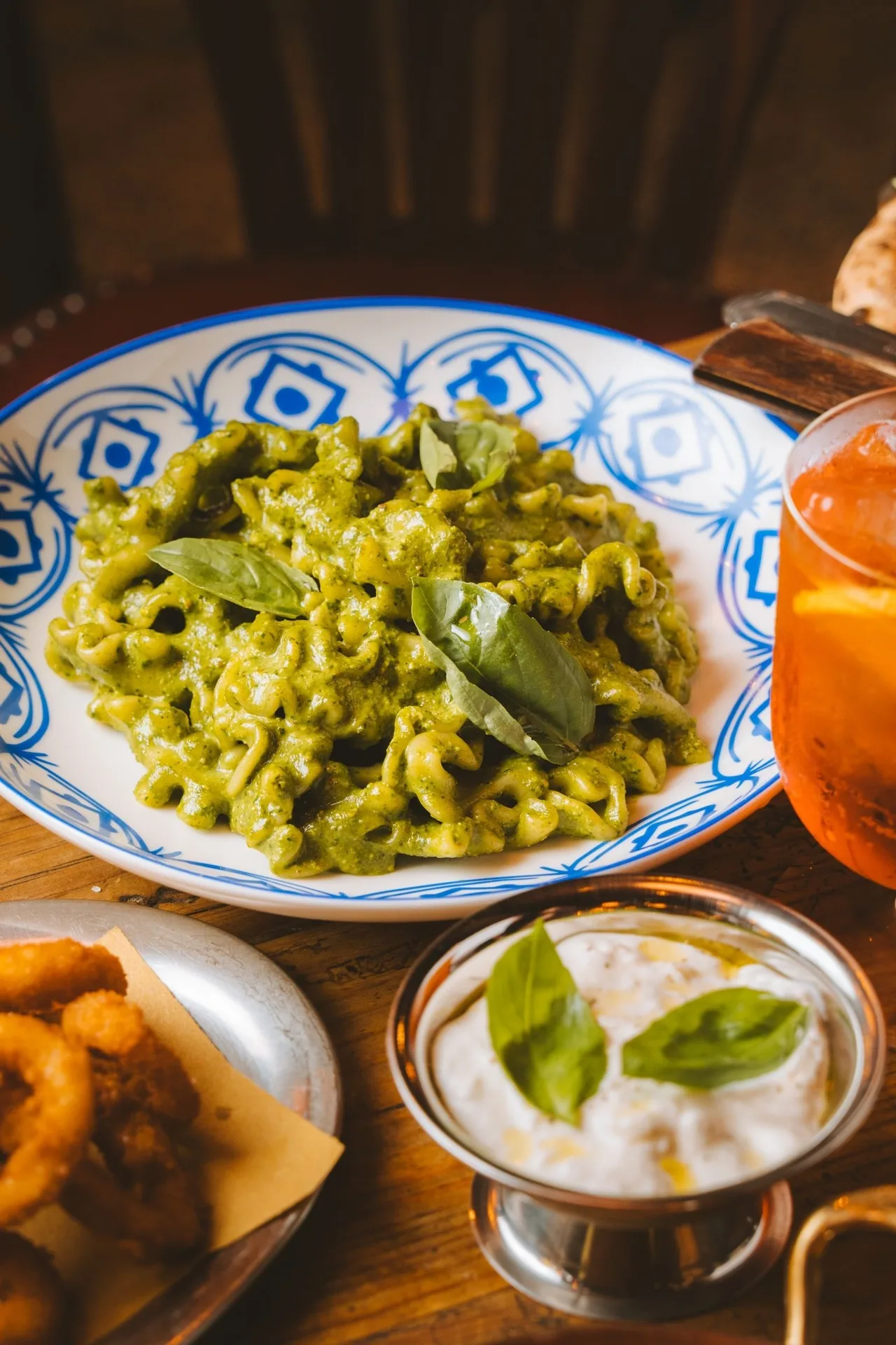 A blue and white patterned plate of green pasta with basil leaves and a bowl of white cream beside it