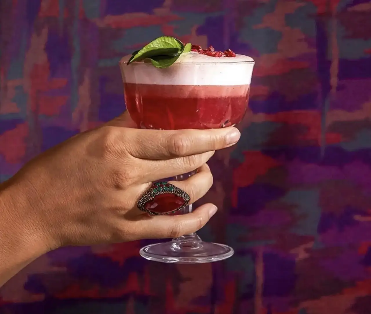 A hand wearing a jewelled ring holds a stemmed glass of layered red cocktail topped with white foam and a basil leaf against a purple-and-red painted wall