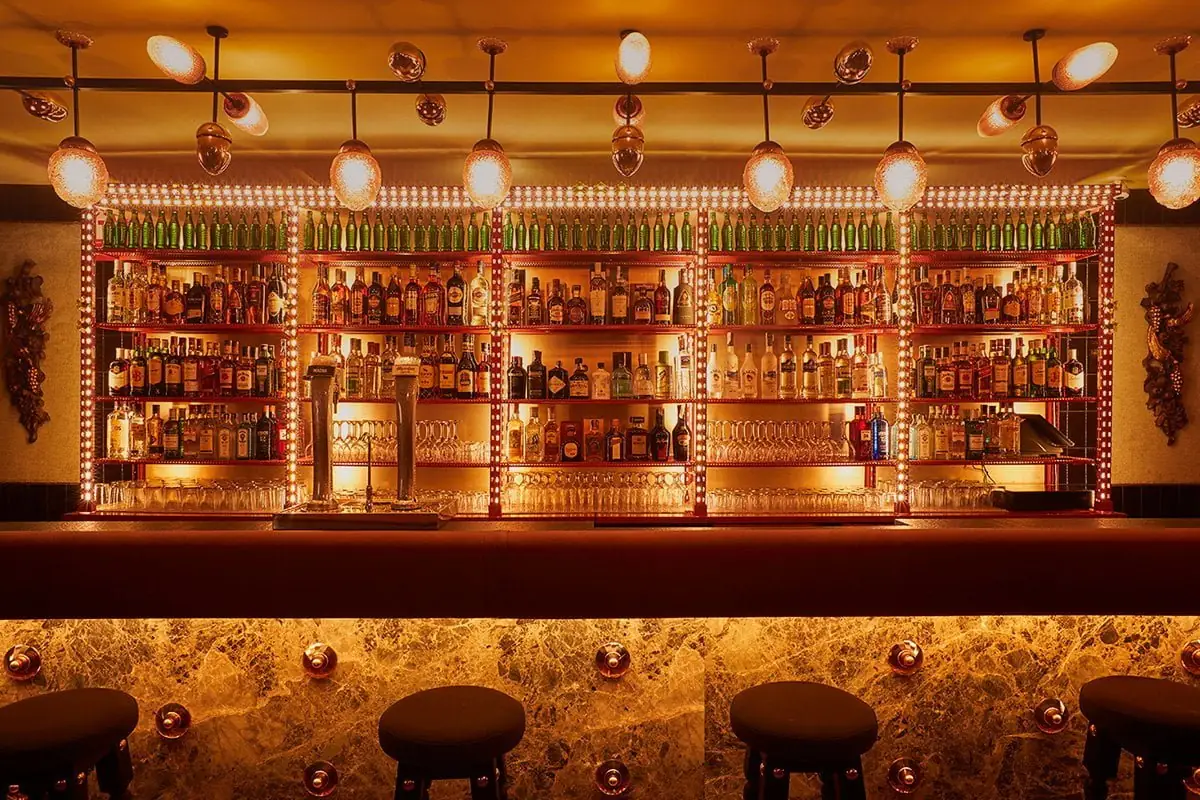 Warmly lit back bar with fairy-light-framed shelving of spirit bottles, green bottles along the top, marble bar front and black stools