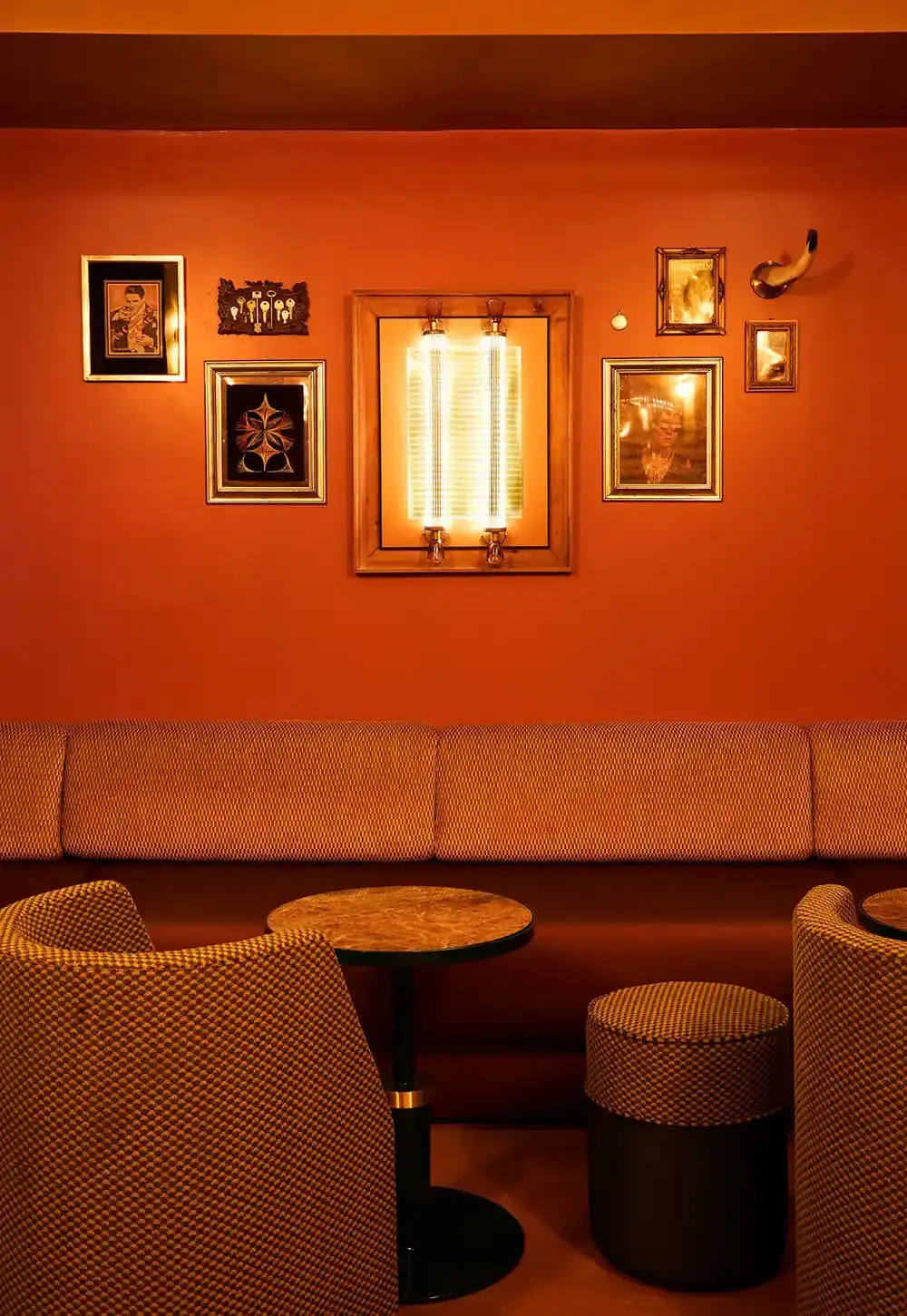 Cosy banquette seating nook with orange walls, framed pictures, an illuminated wall sconce, a round marble table and upholstered tub chairs